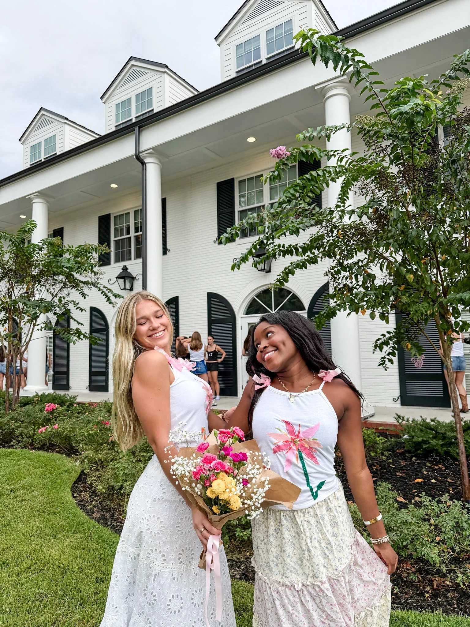 Two women smiling and holding flowers in front of a white house with black shutters and columns, with other people in the background.