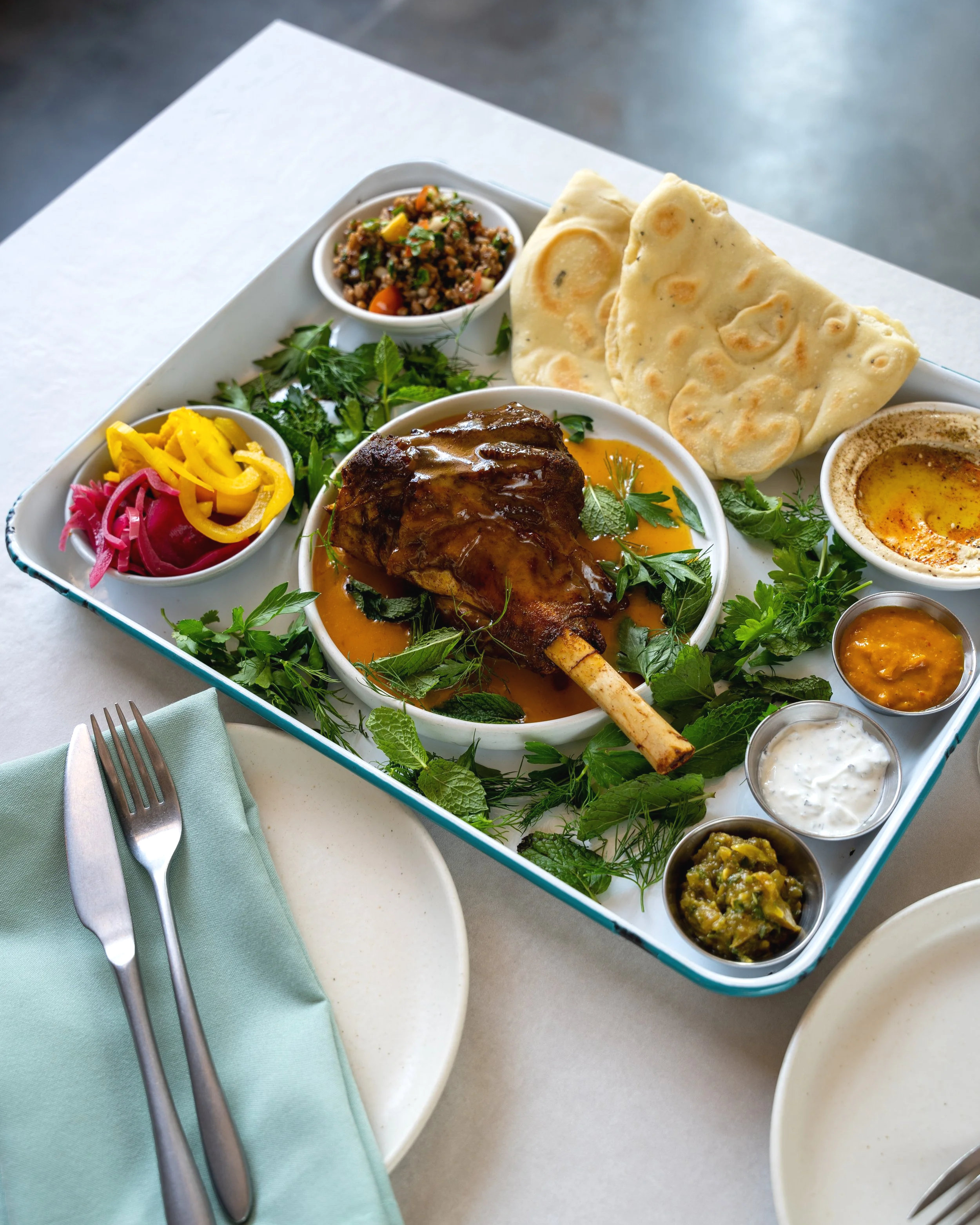 A tray with roasted lamb shank garnished with herbs, pita bread, tabbouleh salad, hummus, and assorted sauces. On the dinner menu at Mayfield restaurant San Juan Capistrano 