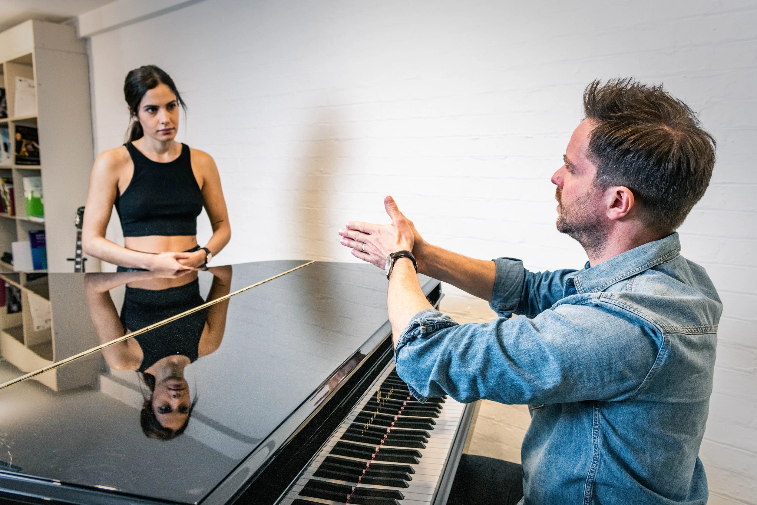 Vocal coach explaining technique to singer beside piano during studio lesson