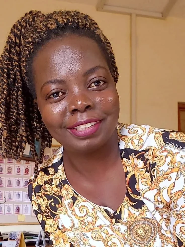 A smiling woman with curly, shoulder-length hair and a patterned shirt, standing in an indoor setting with a bulletin board of photos in the background.