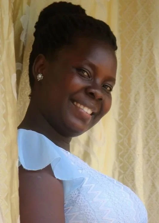 A smiling Ugandan woman  wearing a light blue dress and earrings, standing near a yellow curtain with a patterned background.