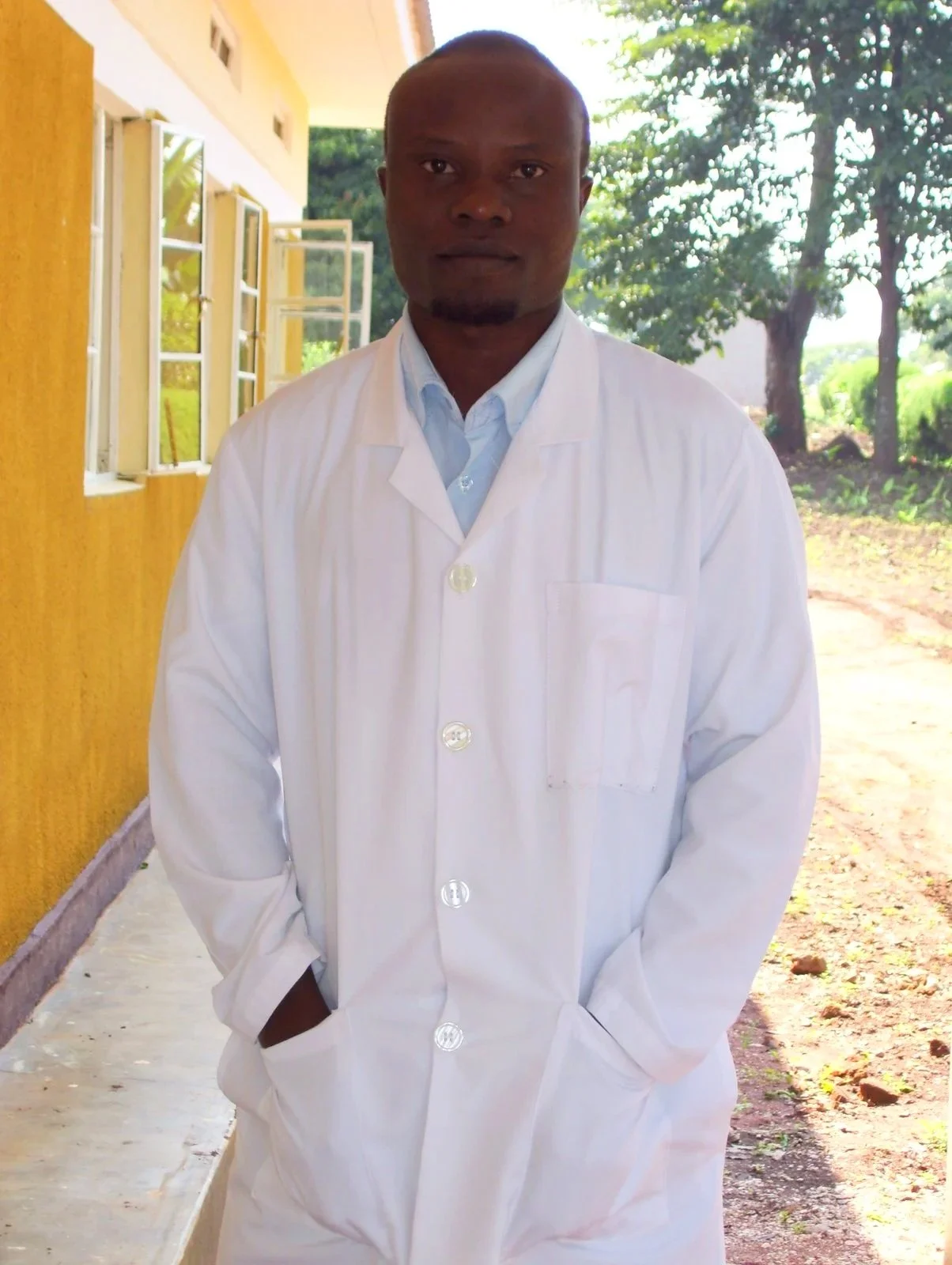A Ugandan male Dr. in a white lab coat standing outside near a yellow building and trees, hands in pockets, looking at the camera.