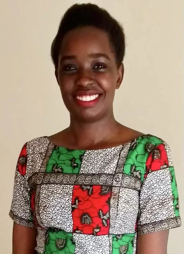 Smiling young Ugandan woman with short hair in a colorful patterned dress standing against a plain background.