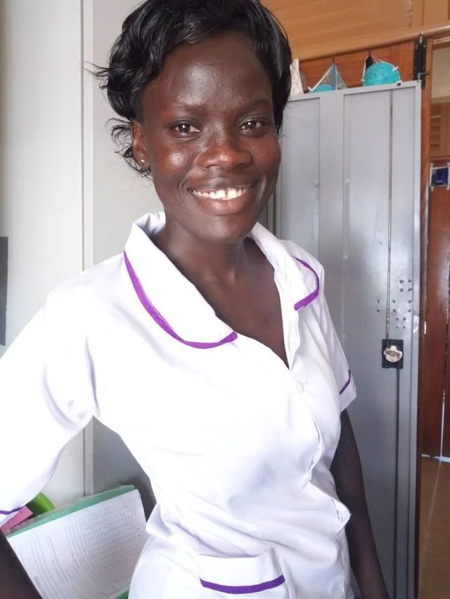A Ugandan woman with short black hair smiling in a nurse uniform with purple accents, standing in a room with a metallic locker behind her.