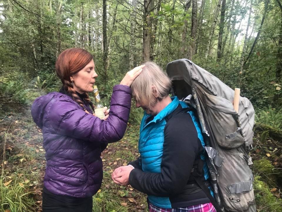 Two women in a forest, one with braided hair and a purple jacket holding an object to the other's forehead, who is wearing a blue jacket and a backpack.