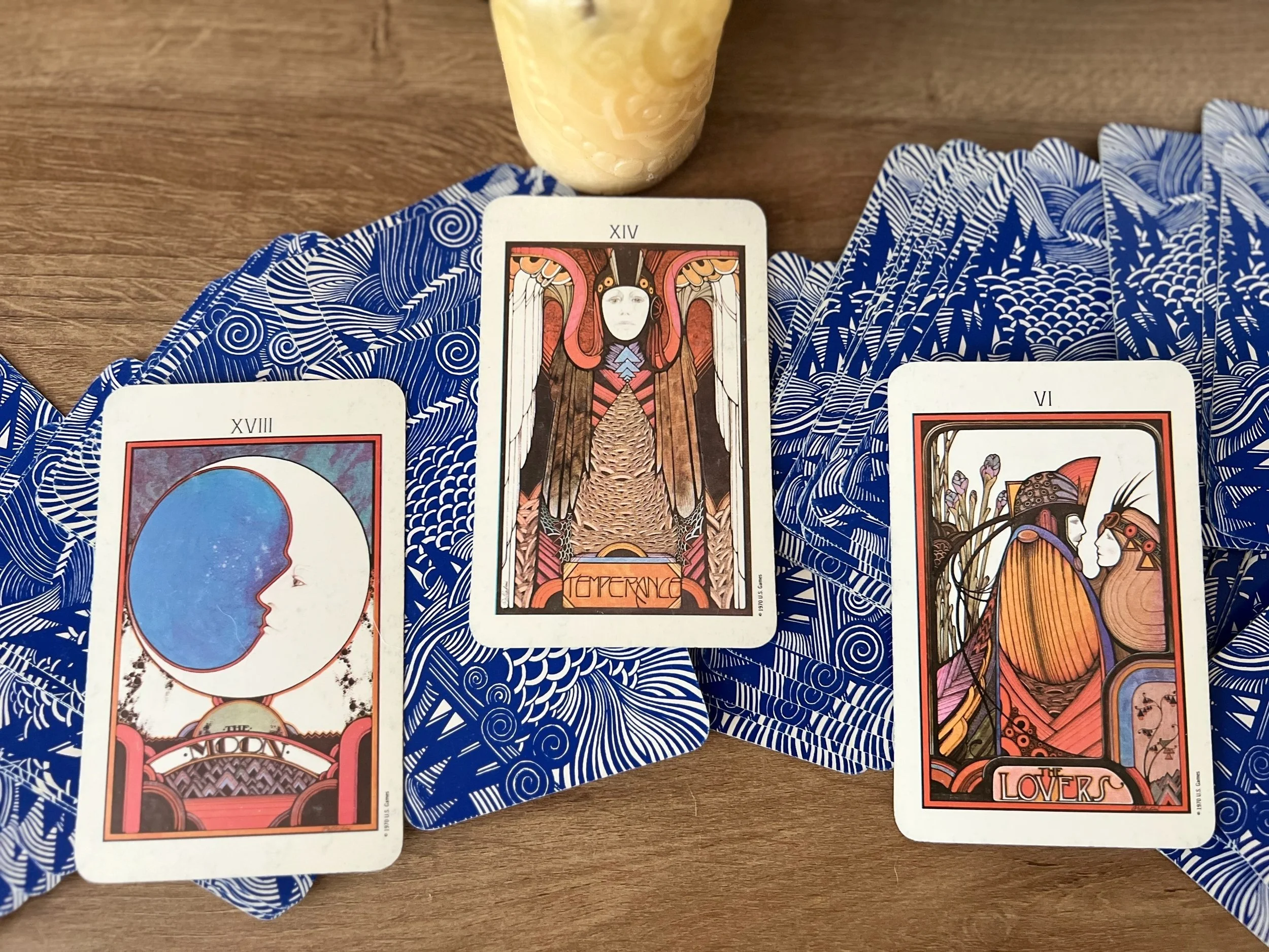 A collection of tarot cards laid out on a blue and white patterned cloth, with a glass of yellow beverage behind them.