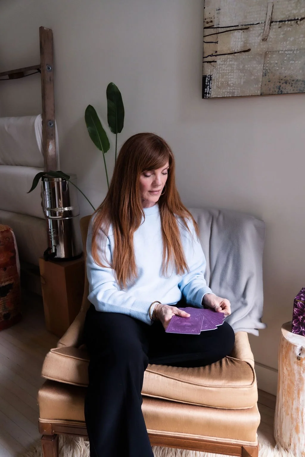 A woman with long red hair wearing a light blue sweater sitting on a beige chair, holding purple tarot cards.