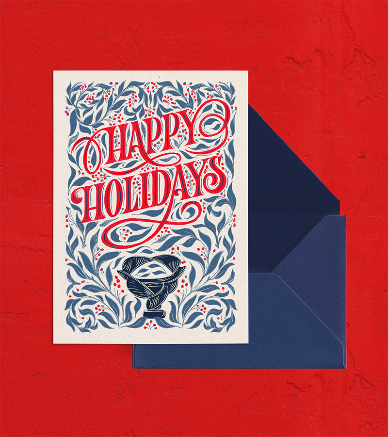 Holiday greeting card with the message "Happy Holidays" in red and blue lettering, surrounded by decorative foliage, and a sculpture illustration at the bottom, placed on a red textured surface with a dark blue envelope.