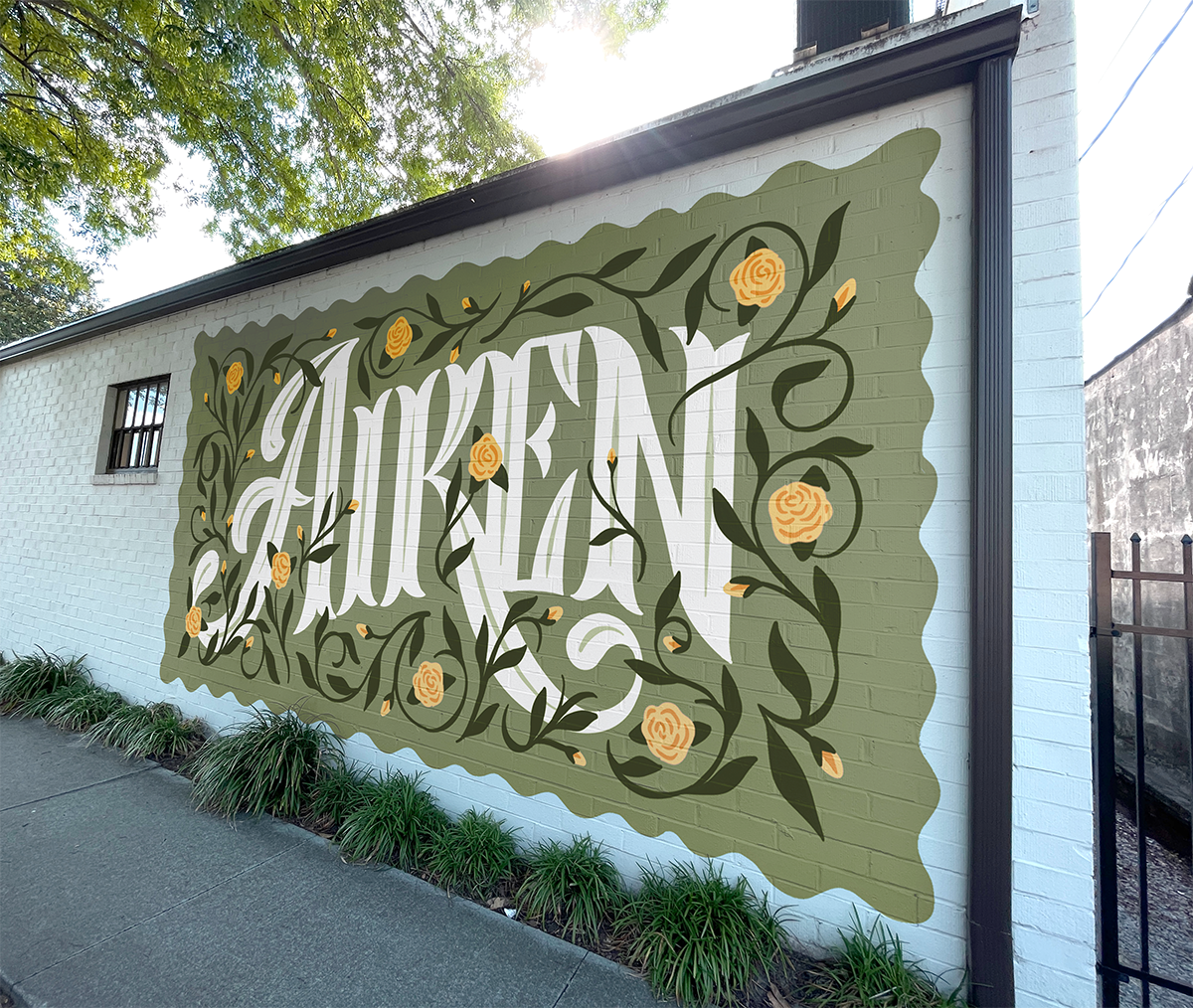 Large mural painted on a white brick wall with the word "Aiken" in stylized white letters surrounded by green vines and yellow roses, with a decorative olive green border.