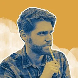 A young male artist with a beard and styled hair, wearing a plaid shirt, is holding a pen contemplatively