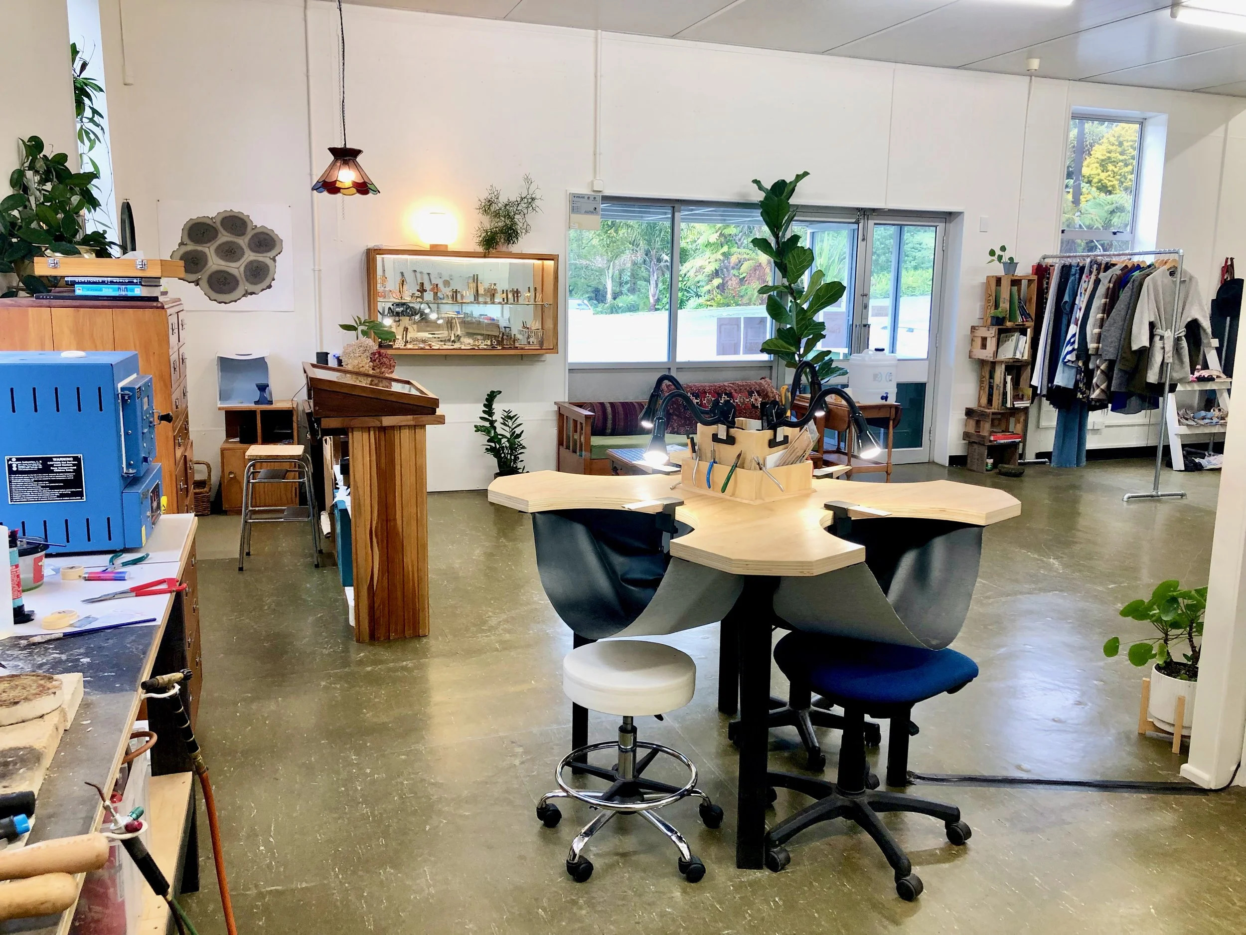 View from workshop space with jewellers bench and tools into the shop space showing counter, jewellery display cabinets and clothing rack in the background.