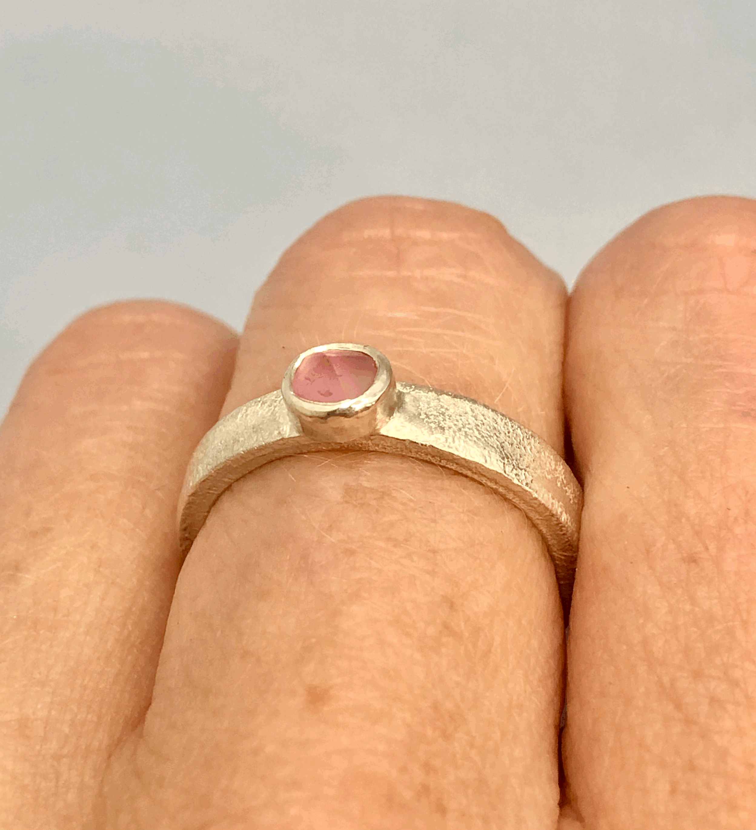 Close-up detail of the pink tourmaline ring worn on finger, highlighting the raw, semi-cut stone texture and the hammered silver band