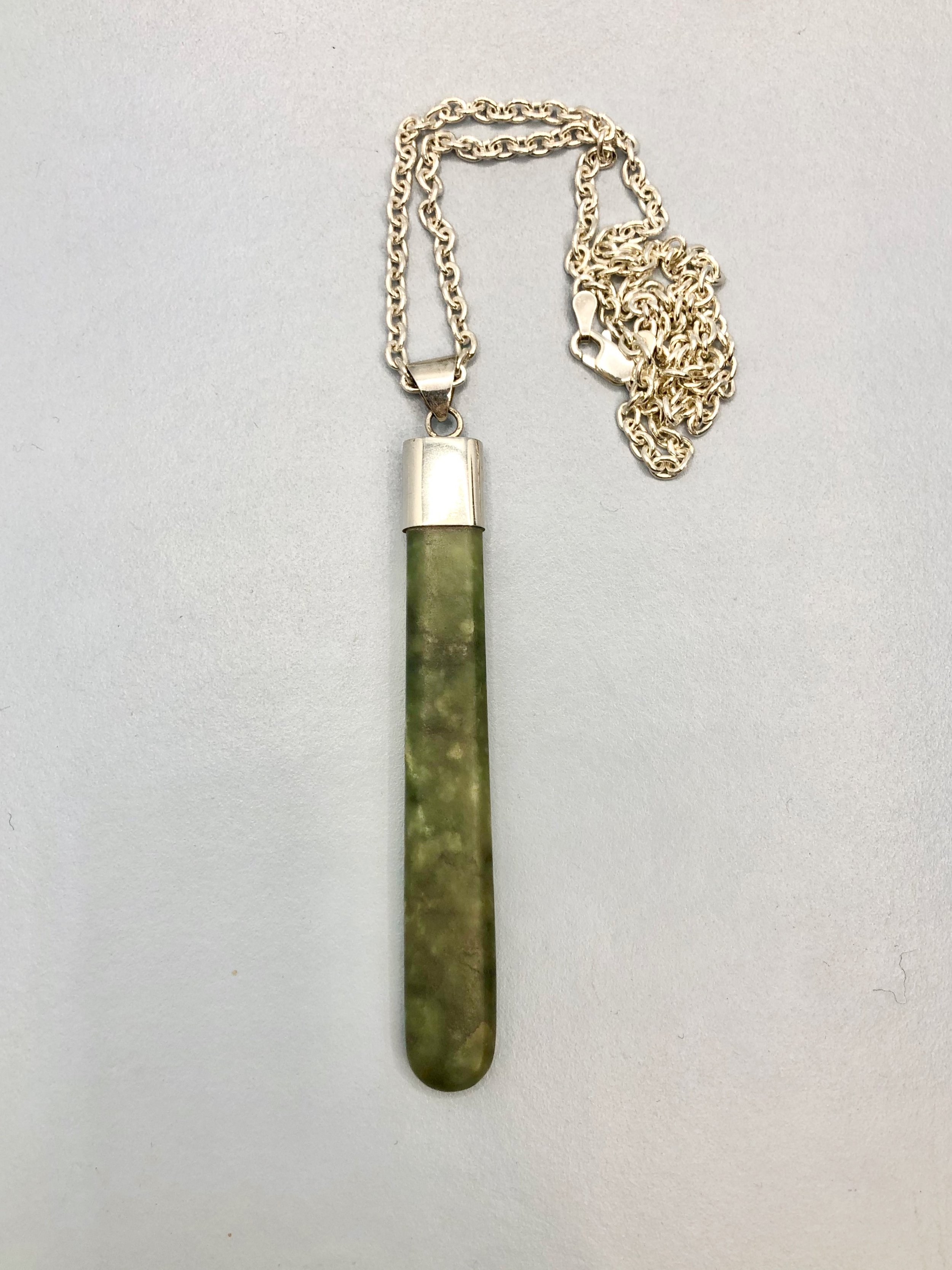 Handmade long pounamu pendant with sterling silver setting lying down front on.