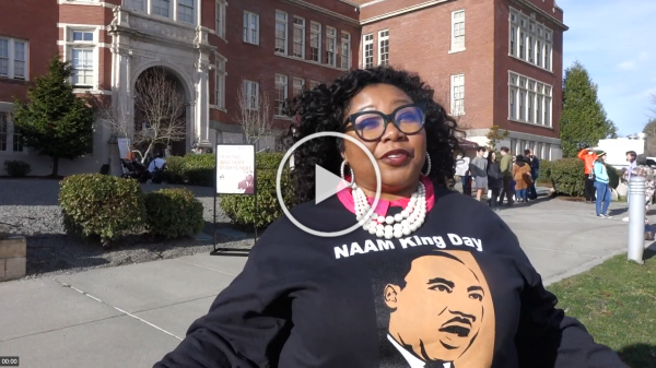 Woman wearing glasses, pearl necklace, and a shirt with Martin Luther King Jr.'s portrait, standing in front of a historic building during a public event.