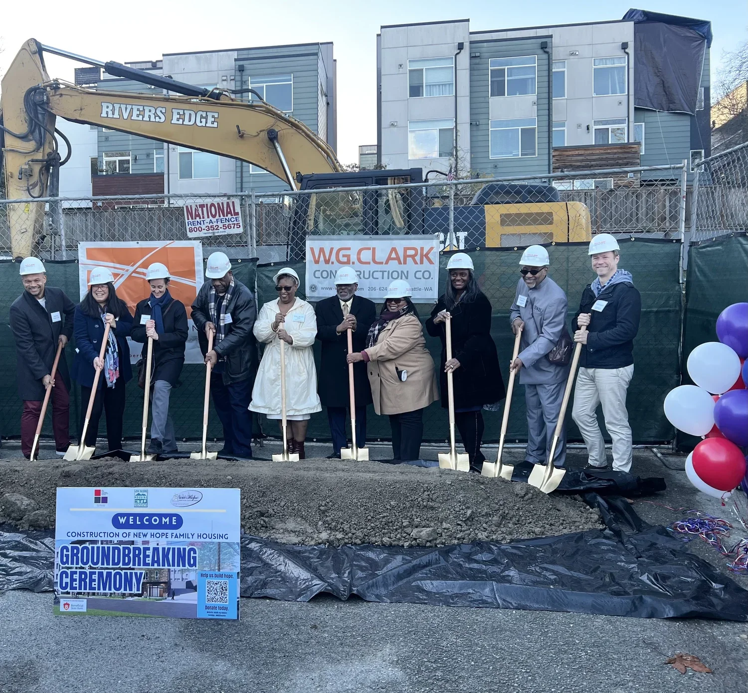 Group of people wearing hard hats at a groundbreaking ceremony for new Hope family housing, holding shovels, standing in front of a sign that says 'Groundbreaking Ceremony' with balloons on the right, in an urban area with modern buildings in the background.