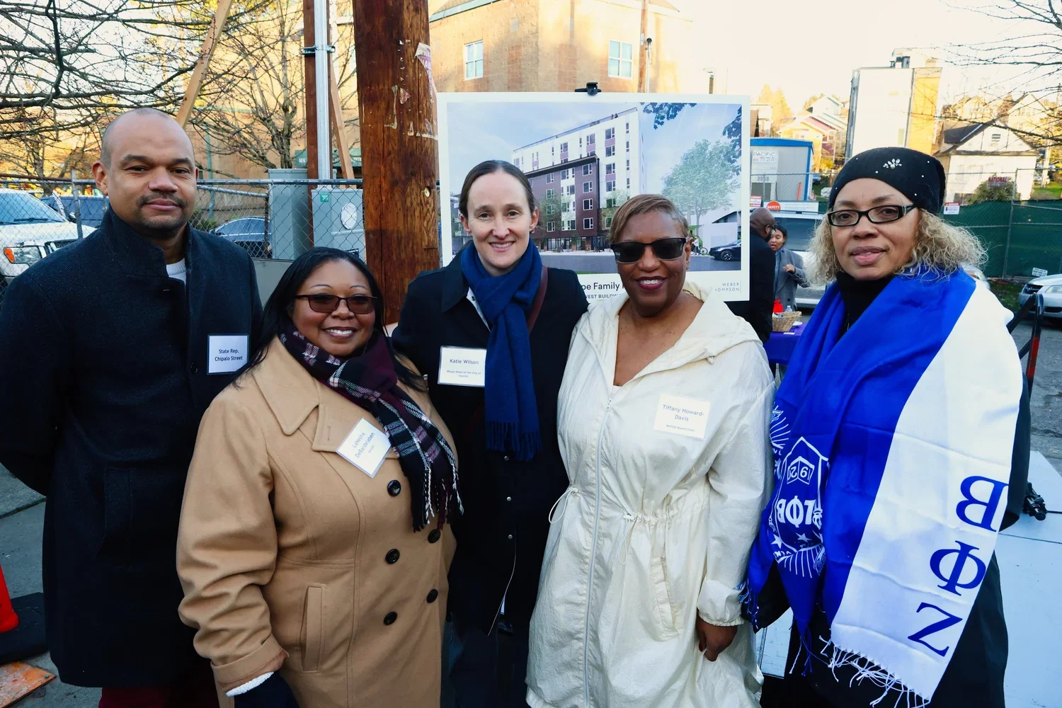 A group of five diverse women and one man standing outdoors, smiling at a public event, with a construction site and parked cars in the background.