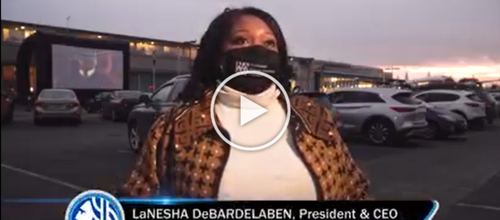 A woman with long black hair, wearing a black mask and patterned jacket, being interviewed outdoors in a parking lot during sunset.