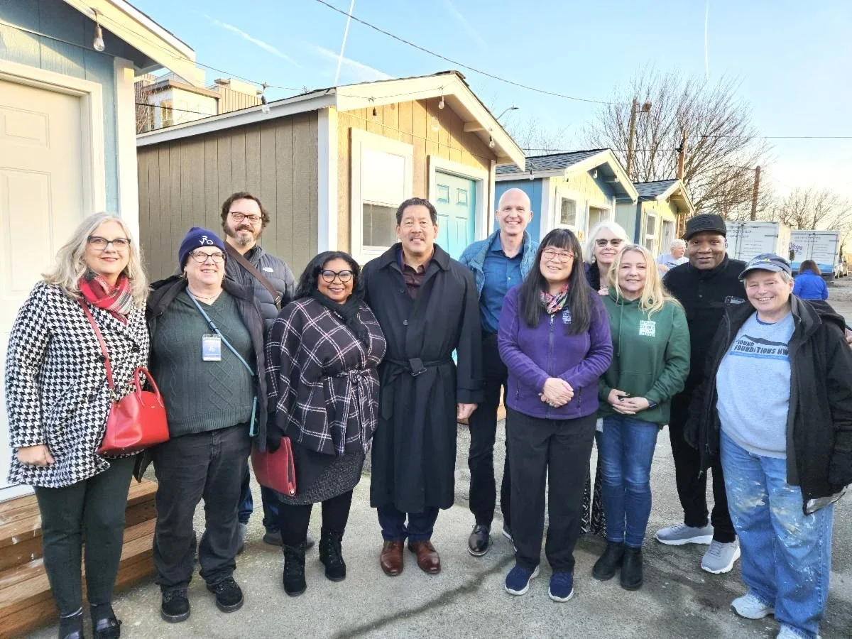 A diverse group of people standing outdoors, smiling for the camera in front of small, colorful houses during daylight.