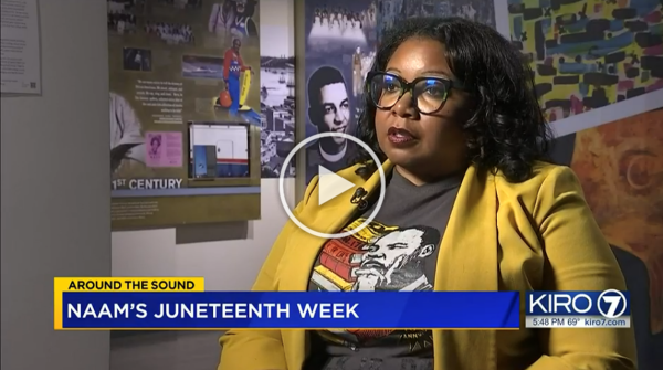 Woman with glasses wearing a yellow blazer and black t-shirt sitting in a room with cultural posters on the wall, being interviewed for a news segment about NAAM's Juneteenth week.