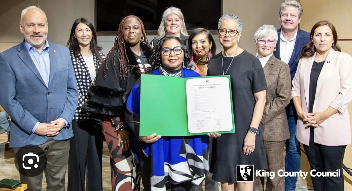 A group of ten diverse people standing in the King County chambers representing King County Council and community holding a Black History proclamation in a green folder.