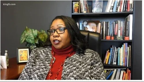A woman with glasses and dark hair speaking in an office, with a bookshelf and a potted plant in the background.