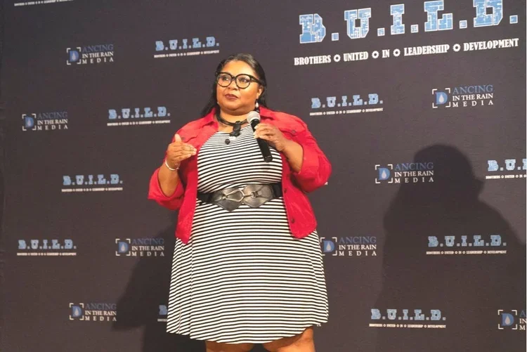 A woman in a black and white striped dress and red jacket speaking into a microphone at a D.O.I.L.D. event on a stage with the D.O.I.L.D. and Dancing in the Rain Media logos on the backdrop.