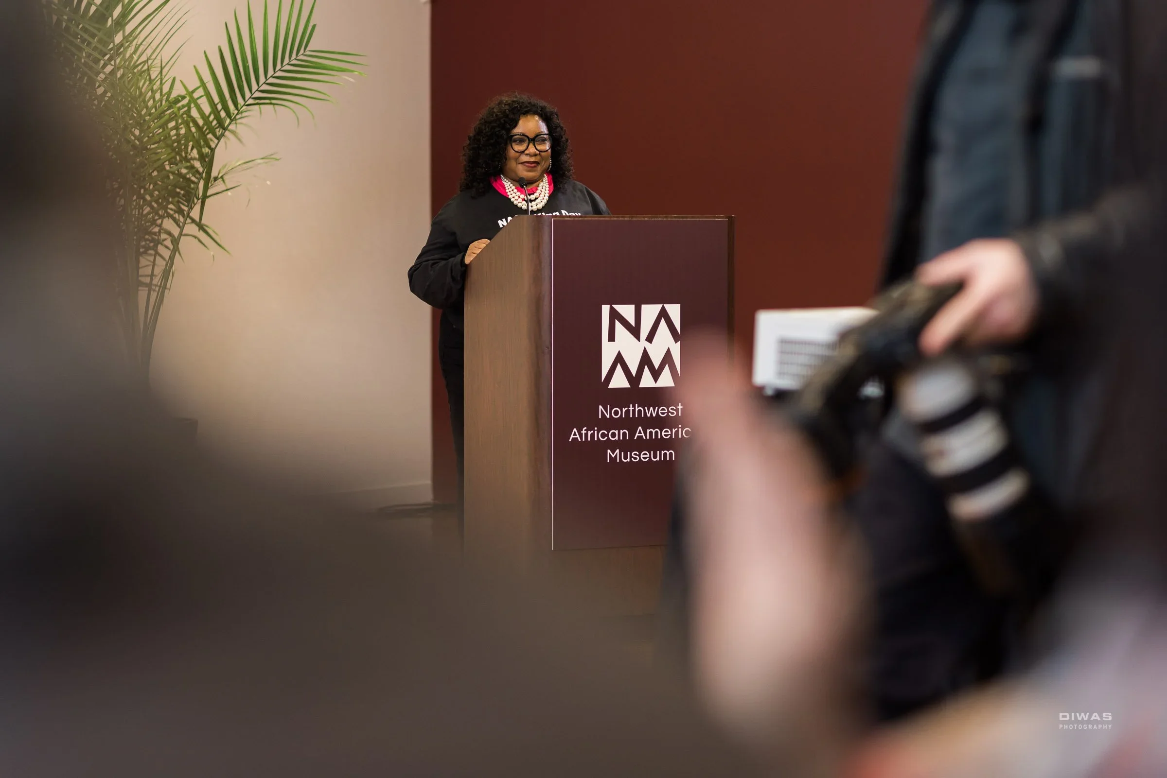 Woman with curly hair, glasses, and pearl necklace speaking at a podium with signage that reads 'Northwest African American Museum'.