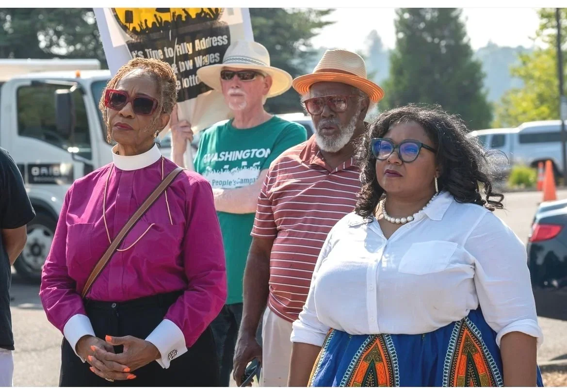 Four diverse individuals standing outdoors during a protest or rally, with cars and trees in the background.
