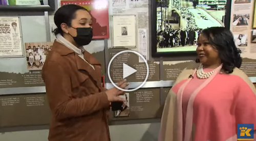 Two women having a conversation indoors, one wearing a black face mask and brown jacket, the other in a pink outfit with a pearl necklace, standing near display boards with historical photos and text.