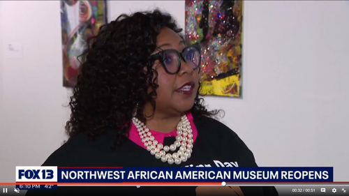 A woman with curly hair and large glasses speaking at an event, wearing a pearl necklace and a black top, in front of colorful artwork.