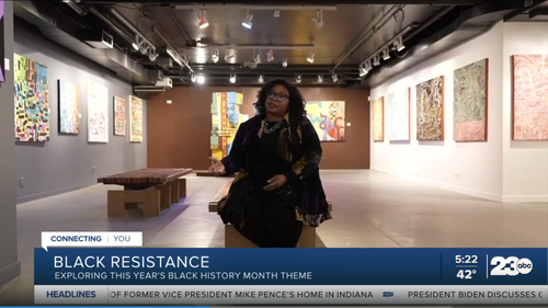A woman with glasses and curly hair is standing in an art gallery with colorful paintings on the walls.