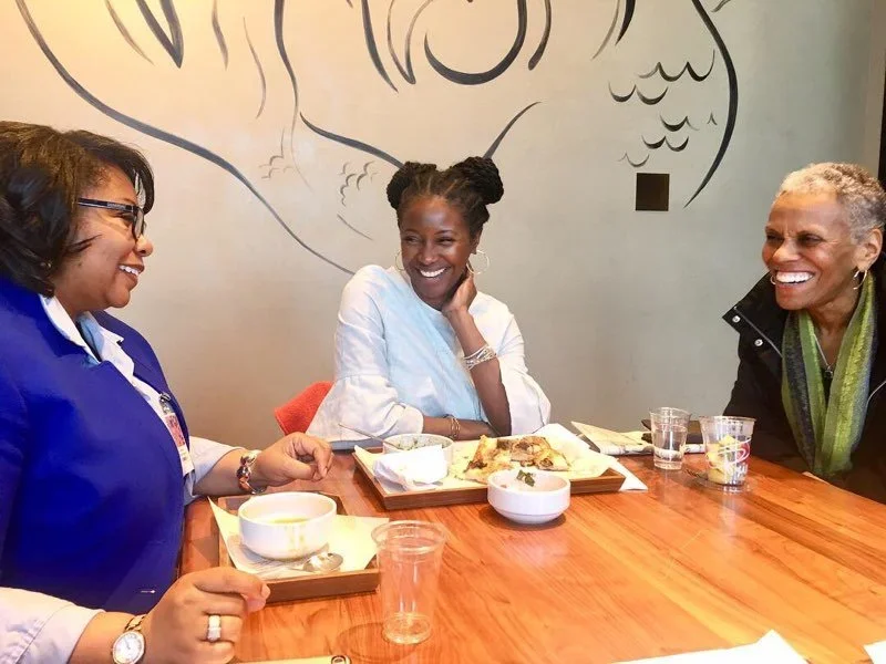 Three women are sitting around a wooden table, smiling and laughing during a meal in a restaurant. The woman on the left wears glasses and a blue outfit, the woman in the middle wears a white blouse, and the woman on the right has short gray hair and wears a black jacket with a green scarf. There are plates with food, bowls, and glasses on the table, and a decorative black line drawing wall art in the background.