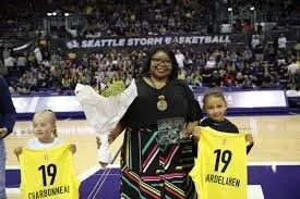 Woman with two young girls on a basketball court during a ceremony or presentation, with the girls holding yellow jerseys numbered 19. The background shows a sports arena with a banner reading 'Seattle Storm & Basketball'.