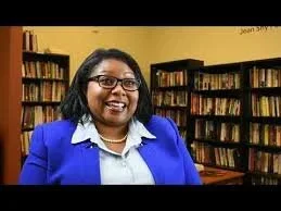A woman with glasses and a blue blazer standing in front of bookshelves in a library.