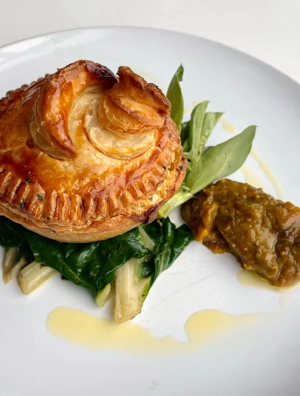 Baked pastry with a spiral design on top, served with greens and a dollop of sauce on a white plate.