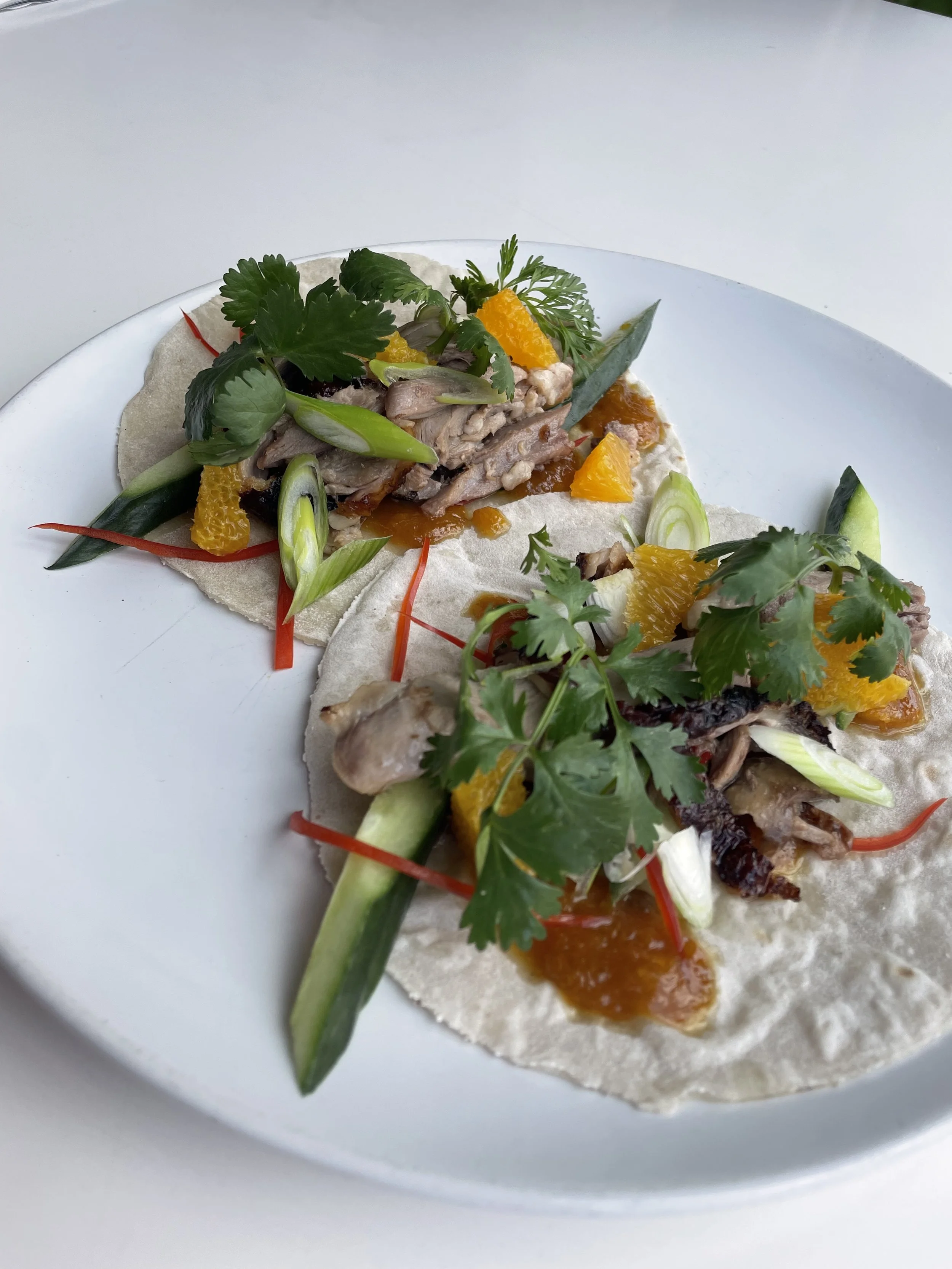 Two tacos with chicken, shredded vegetables, orange slices, cilantro, green onions, and sauce on a white plate.