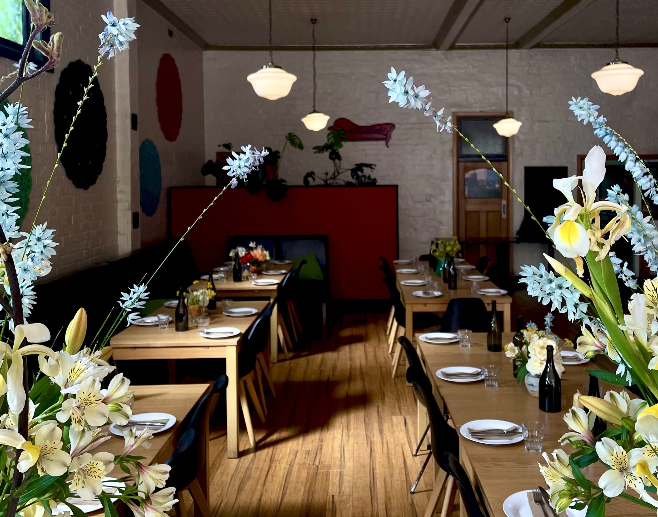 A restaurant interior decorated with floral arrangements and set tables for dining, with hanging ceiling lights and a brick wall.