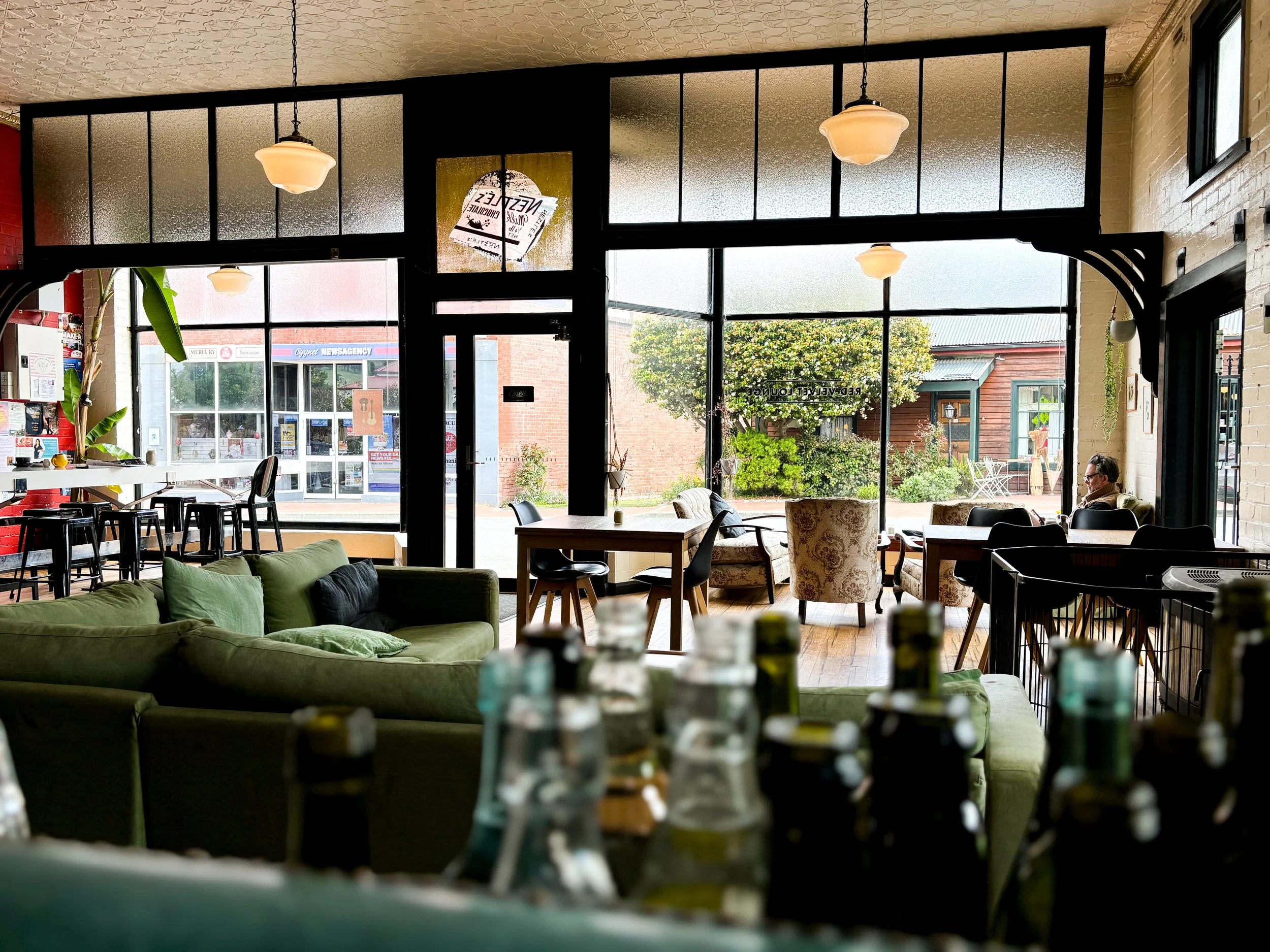Interior of a cozy cafe with a green sofa, armchairs, and tables near large windows showing the outside street and buildings.