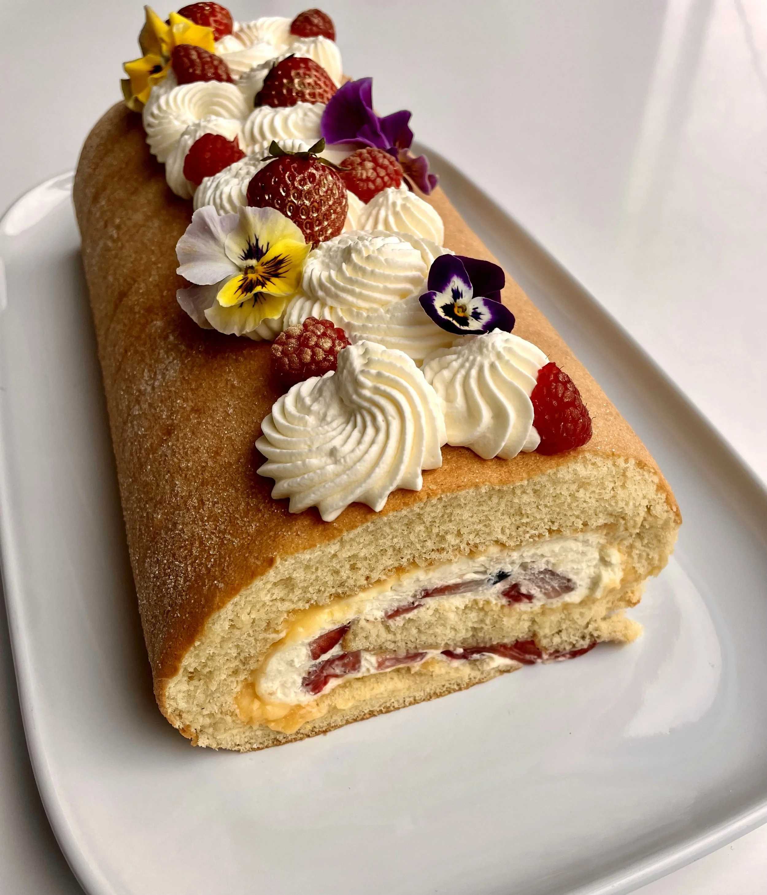 A rolled sponge cake with whipped cream, strawberries, raspberries, and edible flowers on top, served on a white plate.