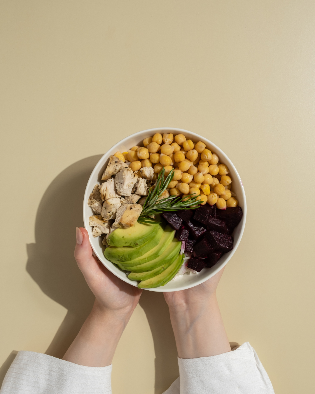 A bowl containing chopped chicken, chickpeas, diced beets, sliced avocado, and a sprig of rosemary, held by two hands against a plain, light-colored background.