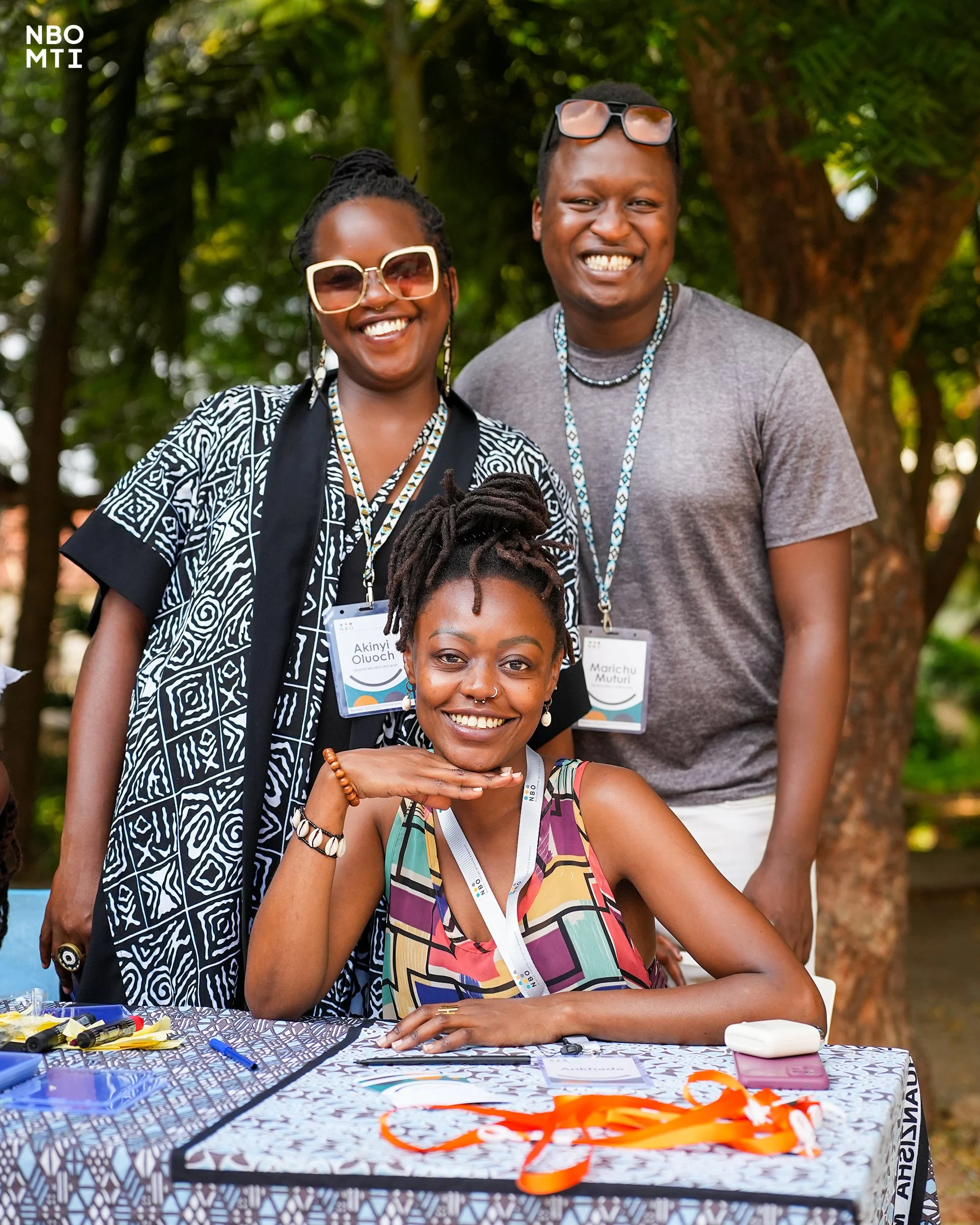 Akinyi, Ankhadada and Marichu pose for a picture looking directly at the camera behind trees at the registration desk at NBO MTI Producing Workshop in Mombasa.