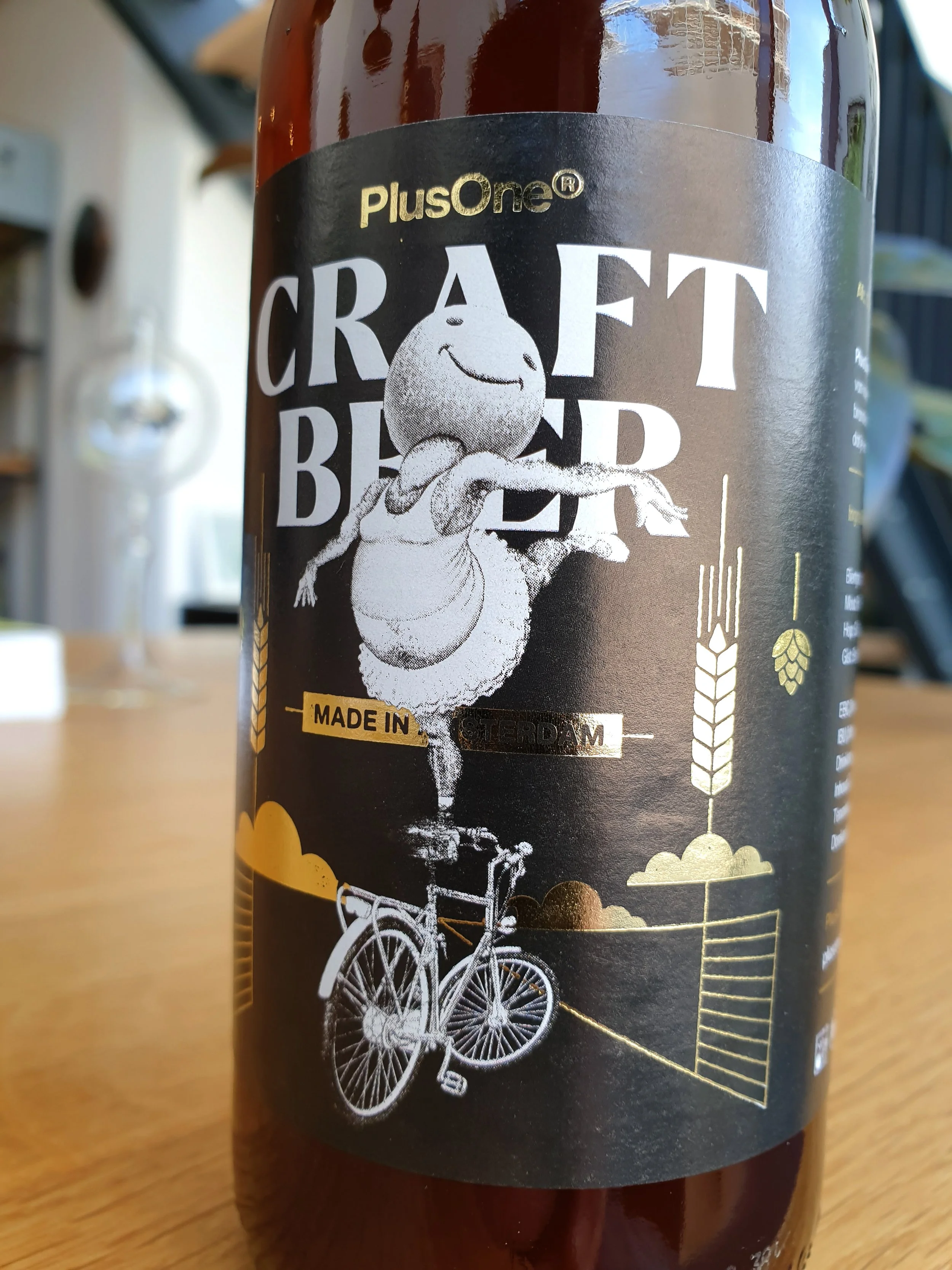 A bottle with a black label featuring a drawing of a happy, smiling cartoon potato riding a vintage bicycle. The label has the text "PlusOne" and "Craft Beer" with some wheat and hop illustrations.