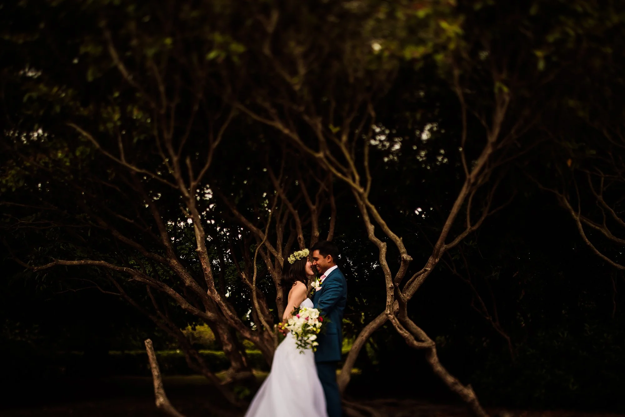A bride and groom standing close together under a tree, smiling, with the bride holding a bouquet of flowers.