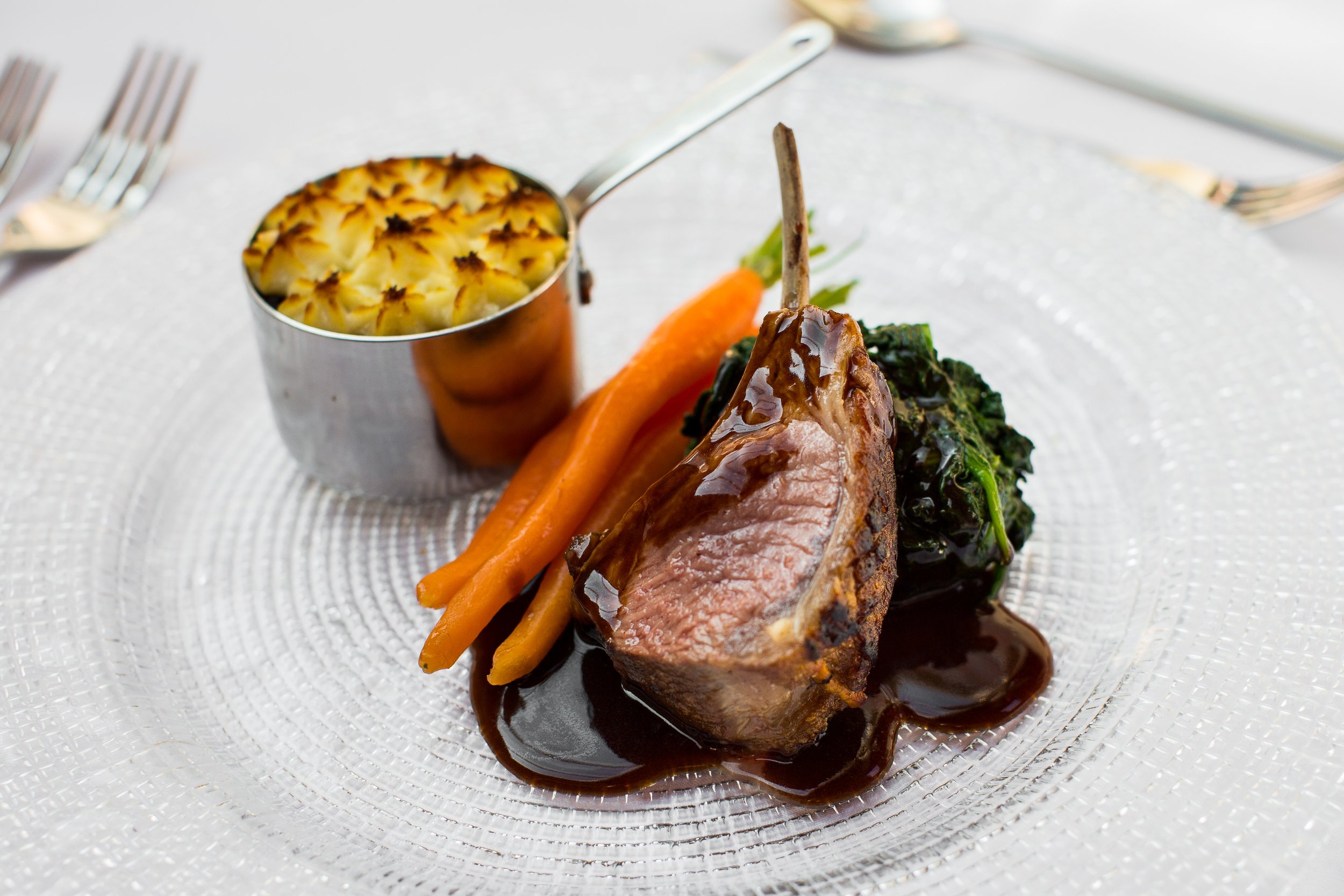 A plated serving of beef tenderloin with dark sauce, garnished with carrots, greens, and roasted garlic, on a textured glass plate.