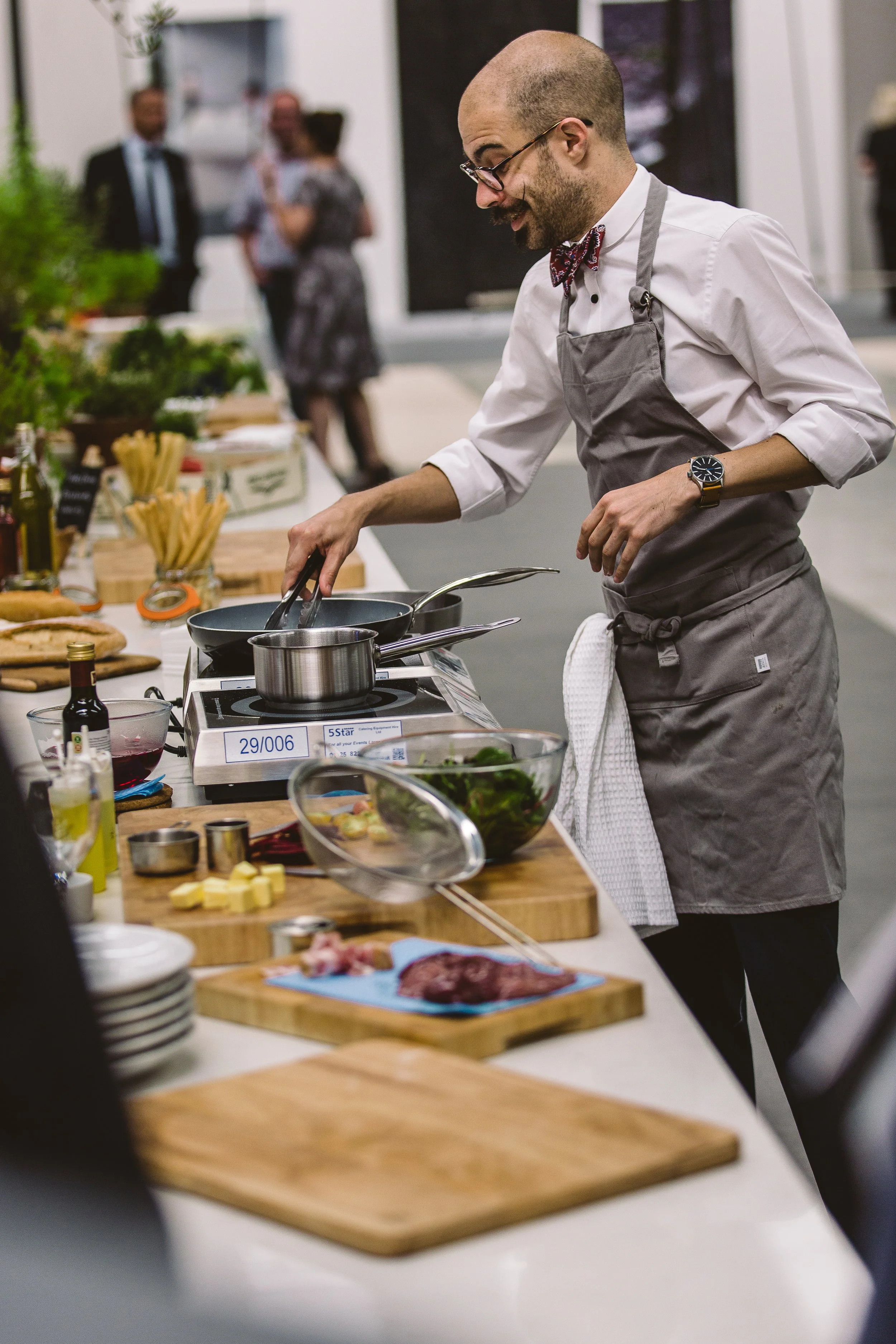 A man with glasses, a beard, and a bow tie is cooking on a portable stove at a kitchen counter with vegetables, herbs, and cooking ingredients around. In the background, there are four people talking and socializing.