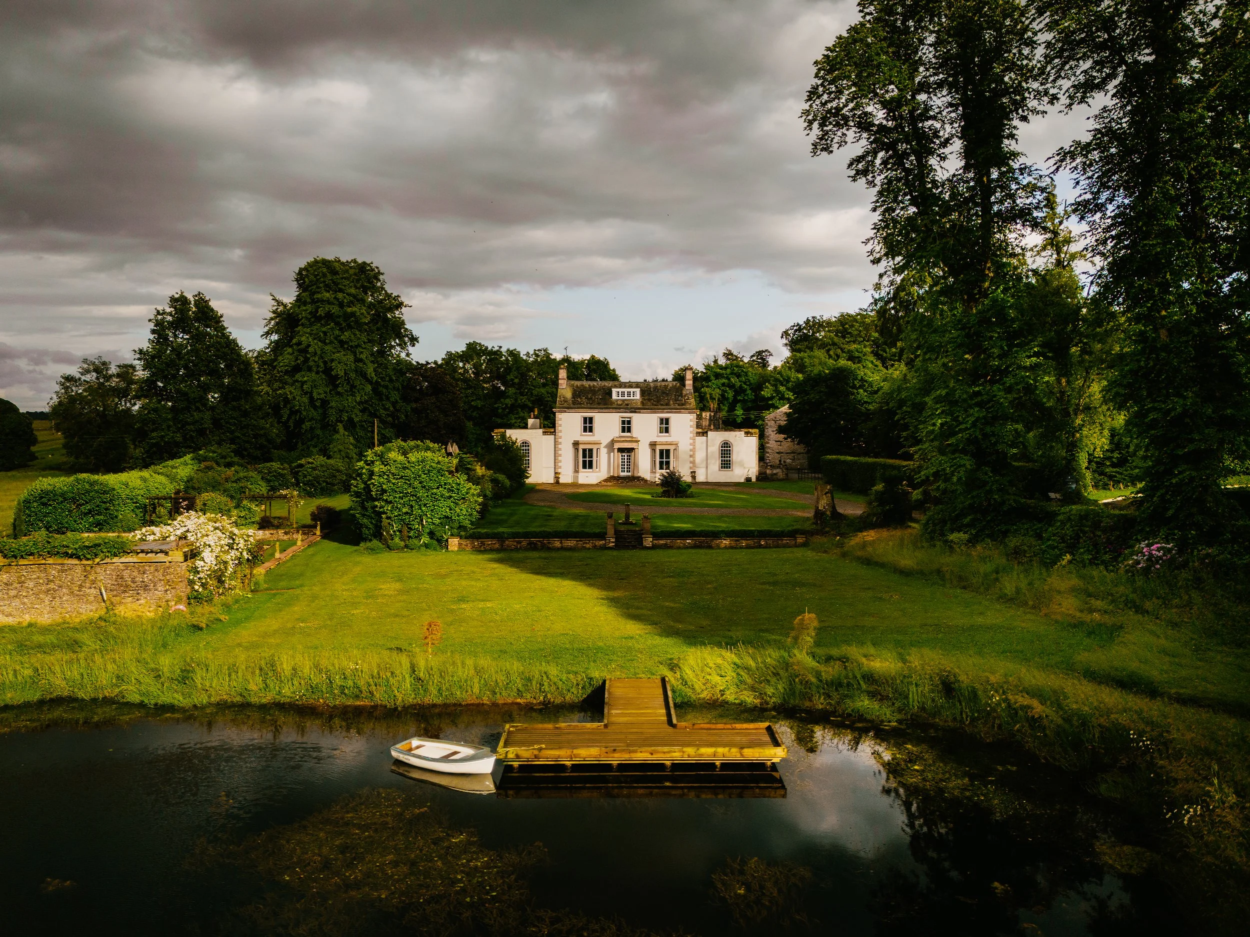 A large white house with a garden, trees, and a dock with a white boat on a calm body of water in the foreground, under a cloudy sky.
