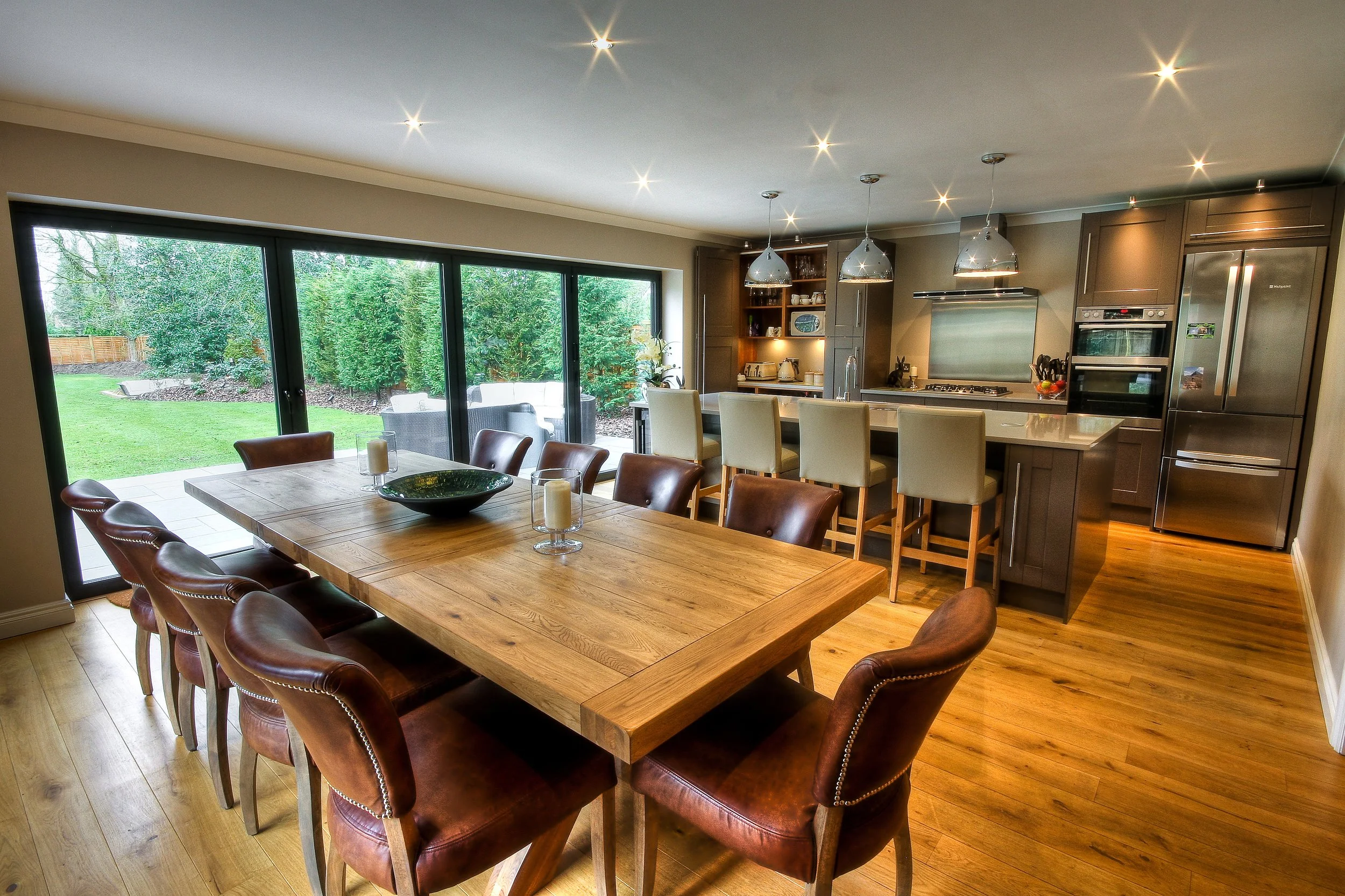 Bright, modern kitchen and dining area with a large wooden table surrounded by brown leather chairs. A sliding glass door opens to a lush green backyard with outdoor furniture.