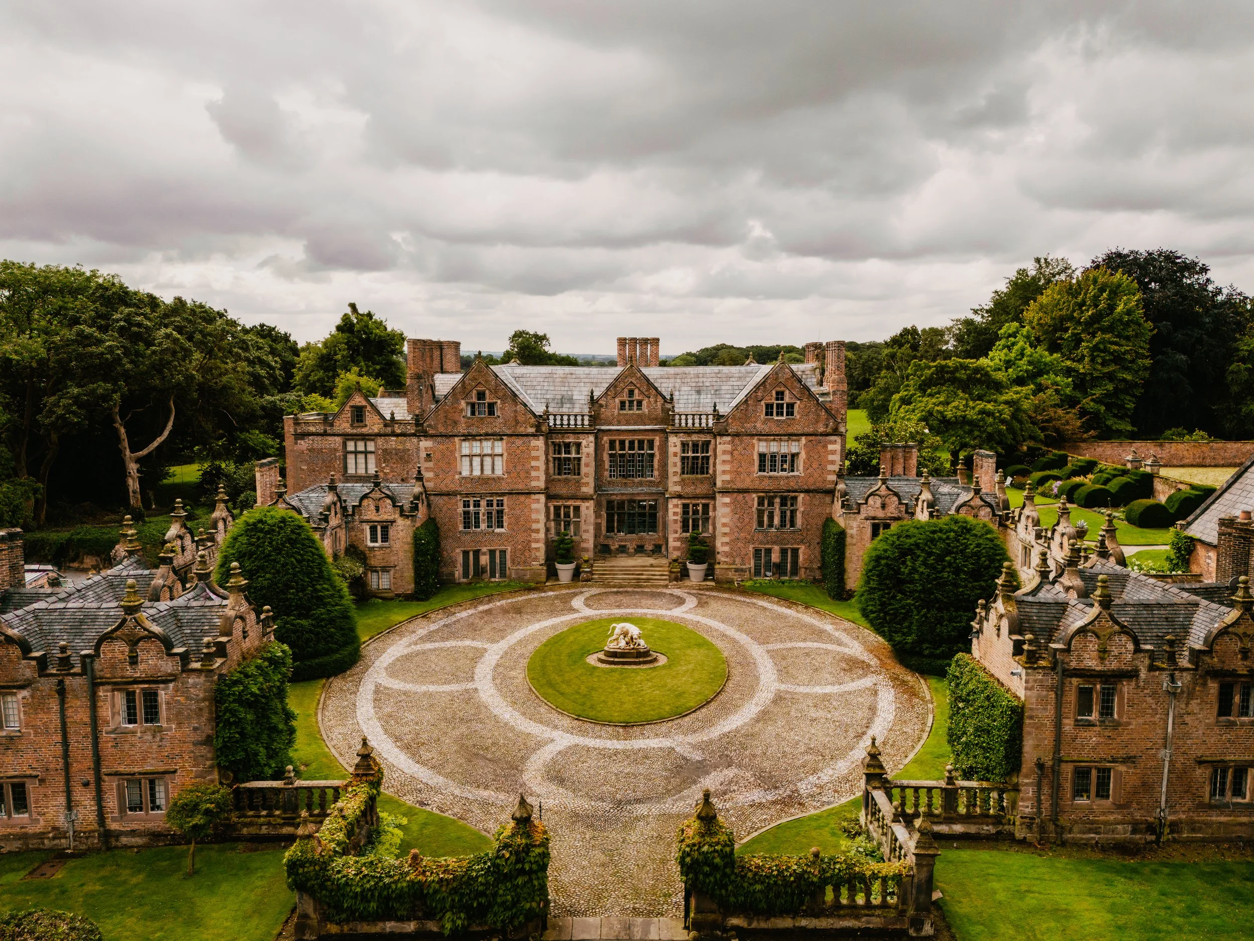 A historic brick mansion with multiple chimneys, overlooking a manicured garden with a circular gravel driveway and a fountain in front. Surrounding trees and a cloudy sky complete the scene.