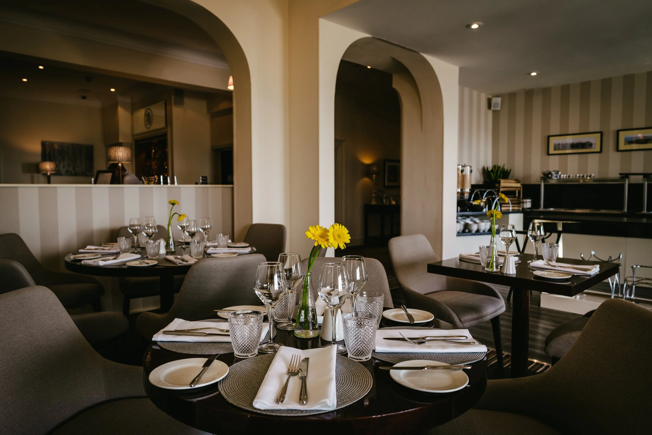 A restaurant dining area with tables set for a meal, including plates, napkins, silverware, and glasses. A small vase with yellow flowers is on each table, and the background shows warm lighting and framed pictures on the wall.