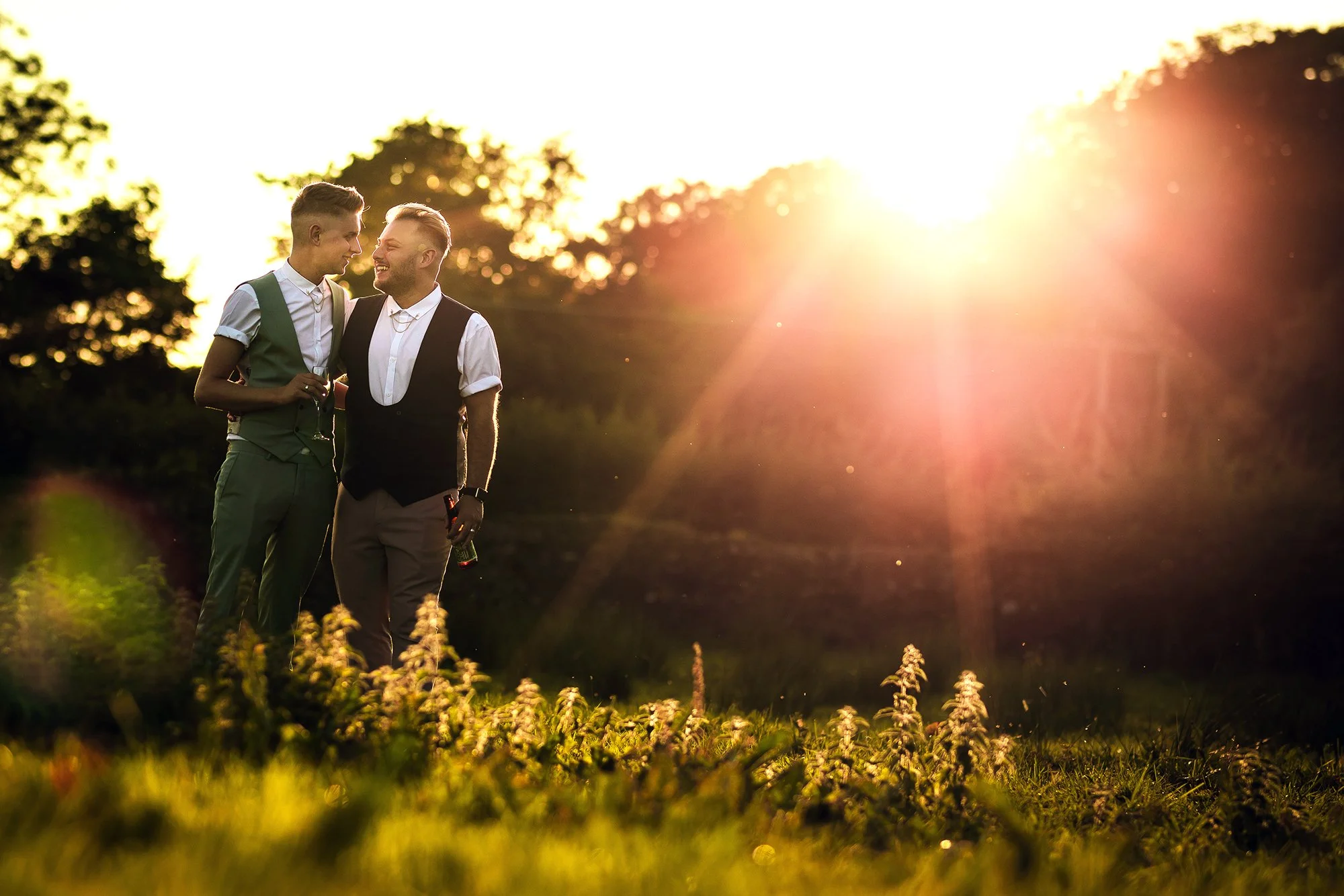 Two men in formal attire standing close together outdoors during sunset, smiling and holding drinks.